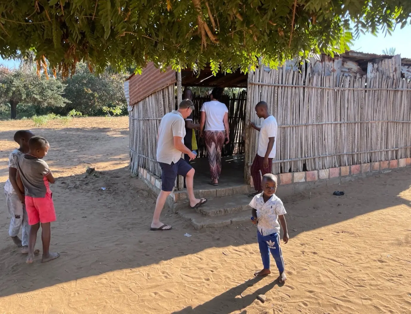 Außenansicht der Schulhütte aus Schilfrohr. Zwei Besucher gehen hinein, Kinder stehen im Sand davor.
