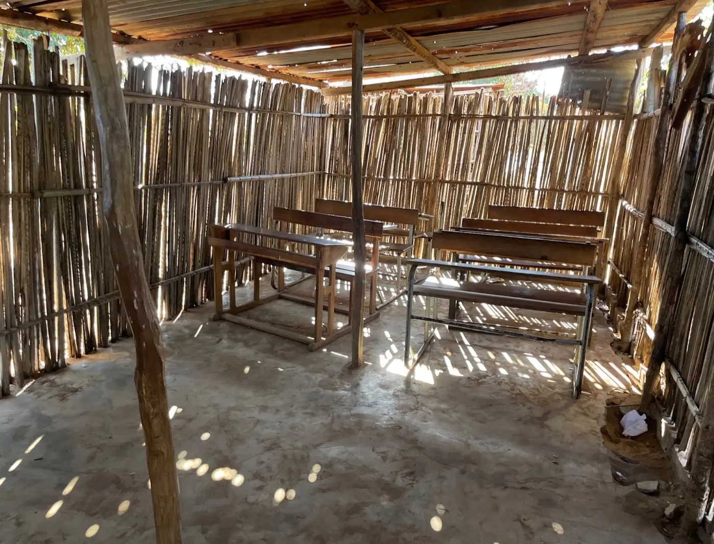 Klassenraum aus Schilfrohrwänden mit Wellblechdach. Durch die Wände fällt Sonnenlicht auf den Betonboden, im Raum stehen einfache Holzbänke.