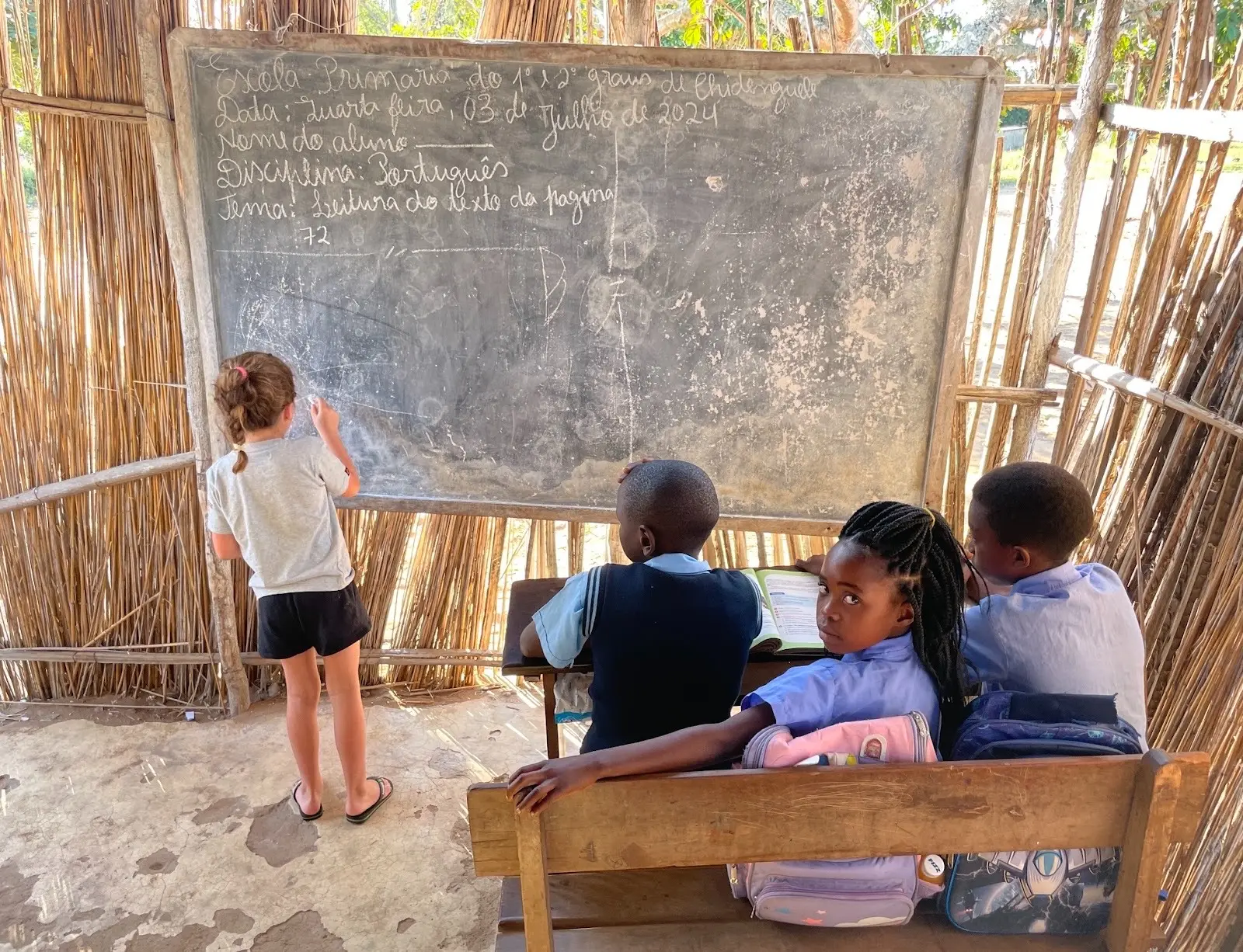 Tafel in einer Schulhütte aus Schilfrohr, beschriftet mit Escola Primária do 1° e 2° graus de Chidenguele, Datum 3. Juli 2024, Fach Portugiesisch. Eine Besucherin schreibt an die Tafel, Kinder schauen zu.